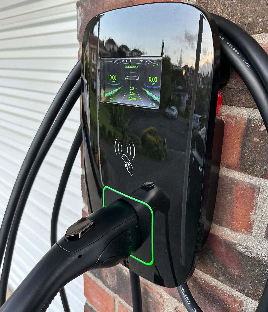 Cargador de pared instalado sobre una pared de ladrillo, con la franja verde encendida y el conector colgado